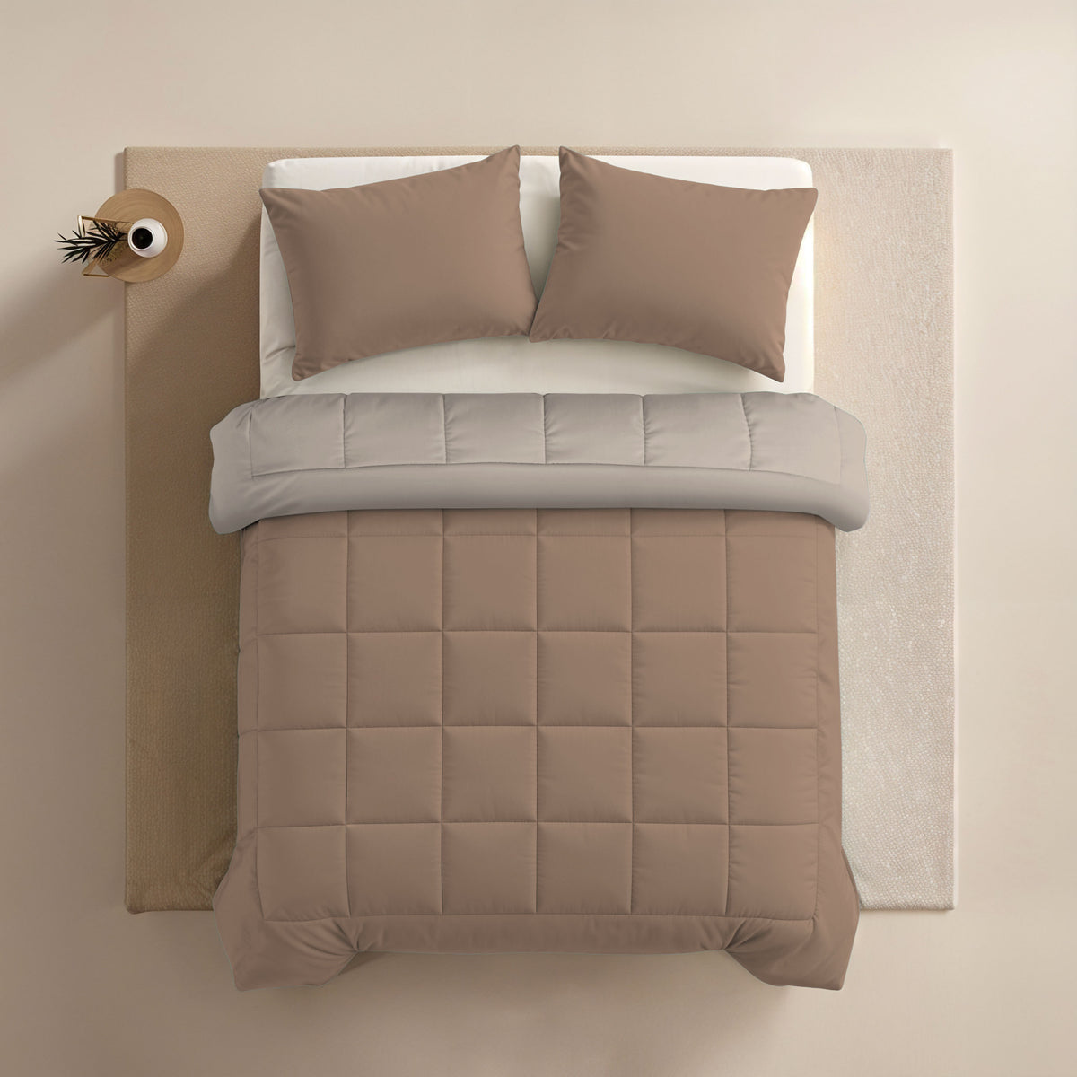 Een netjes opgemaakt bed met een 60x70cm microvezel hoofdkussen met de Zelesta Comfort Duo Kussensloop - Terra, een doorgestikt taupe dekbed, lichtgrijs laken en een klein rond houten bijzettafeltje versierd met een accentstuk.