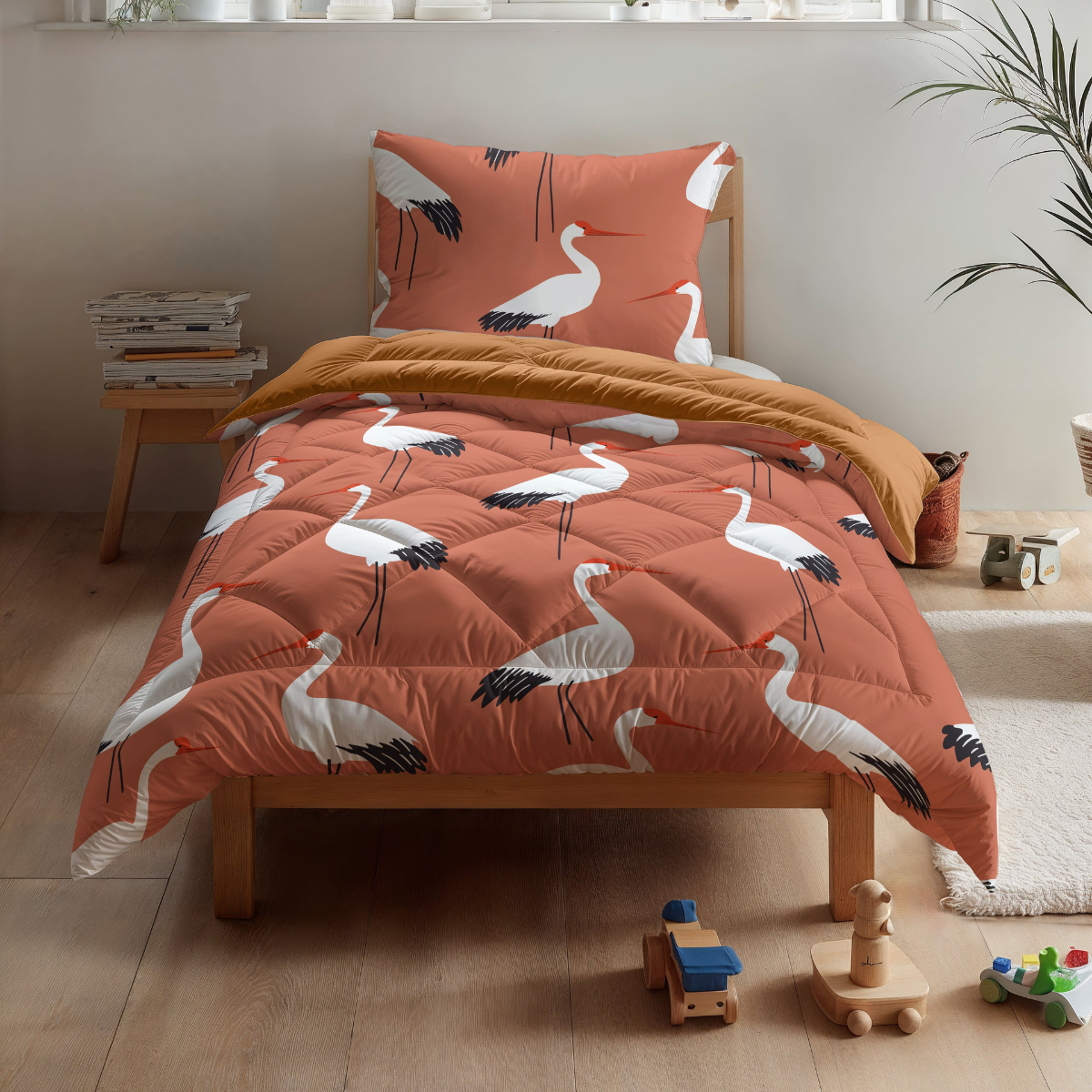 Het Zelesta Kiddybed Katoen - Terra Crane heeft oranje beddengoed met kraanvogelprint op een houten bed, in een lichte kinderkamer met houten vloeren en verspreid liggend speelgoed.