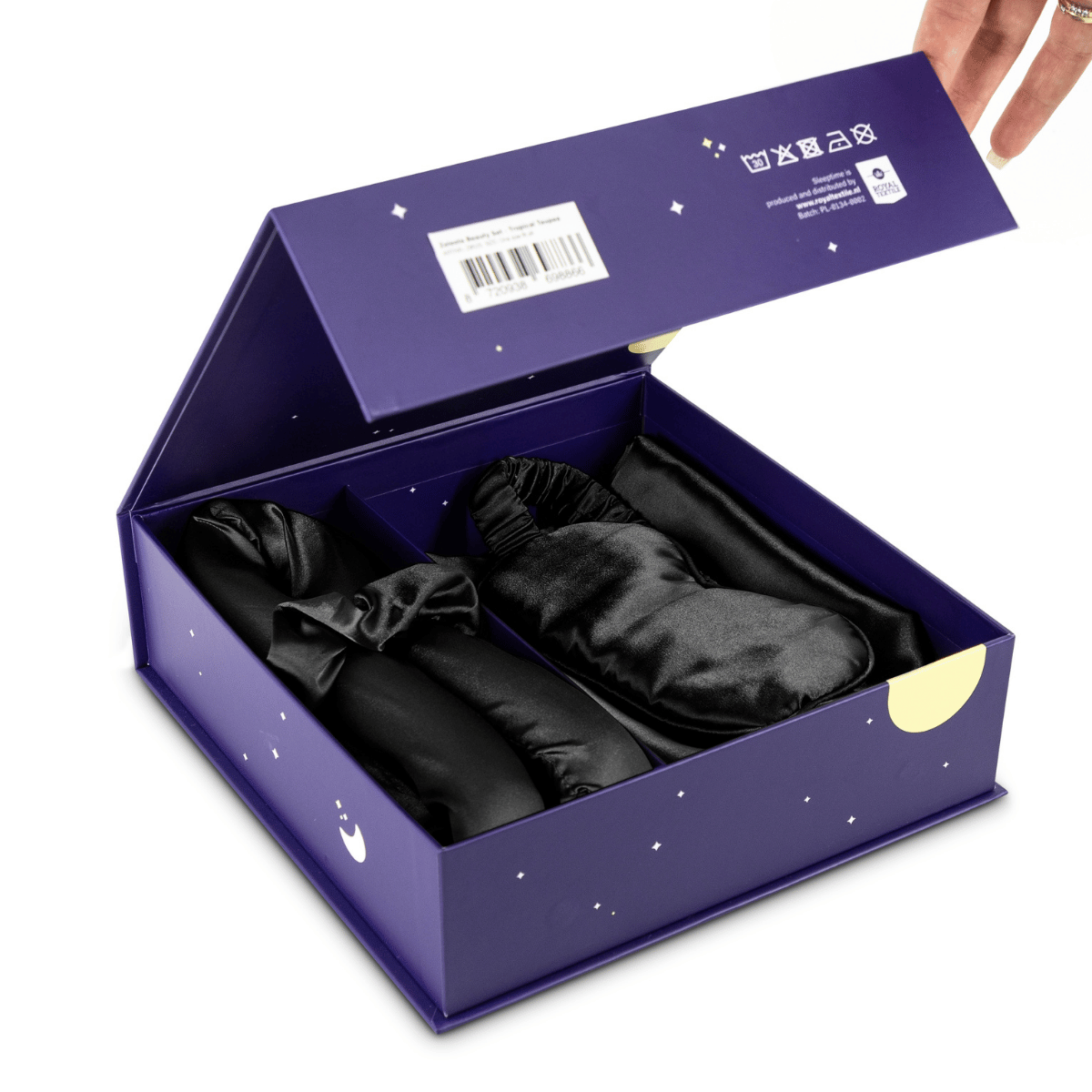 Een hand opent een Zelesta Sleeping Beauty Set - Universal Black geschenkdoos met drie zwart satijnen artikelen, waaronder een kussensloop van moerbeizijde, elk verpakt in zwart - ideaal voor een anti-verouderende slaaproutine.