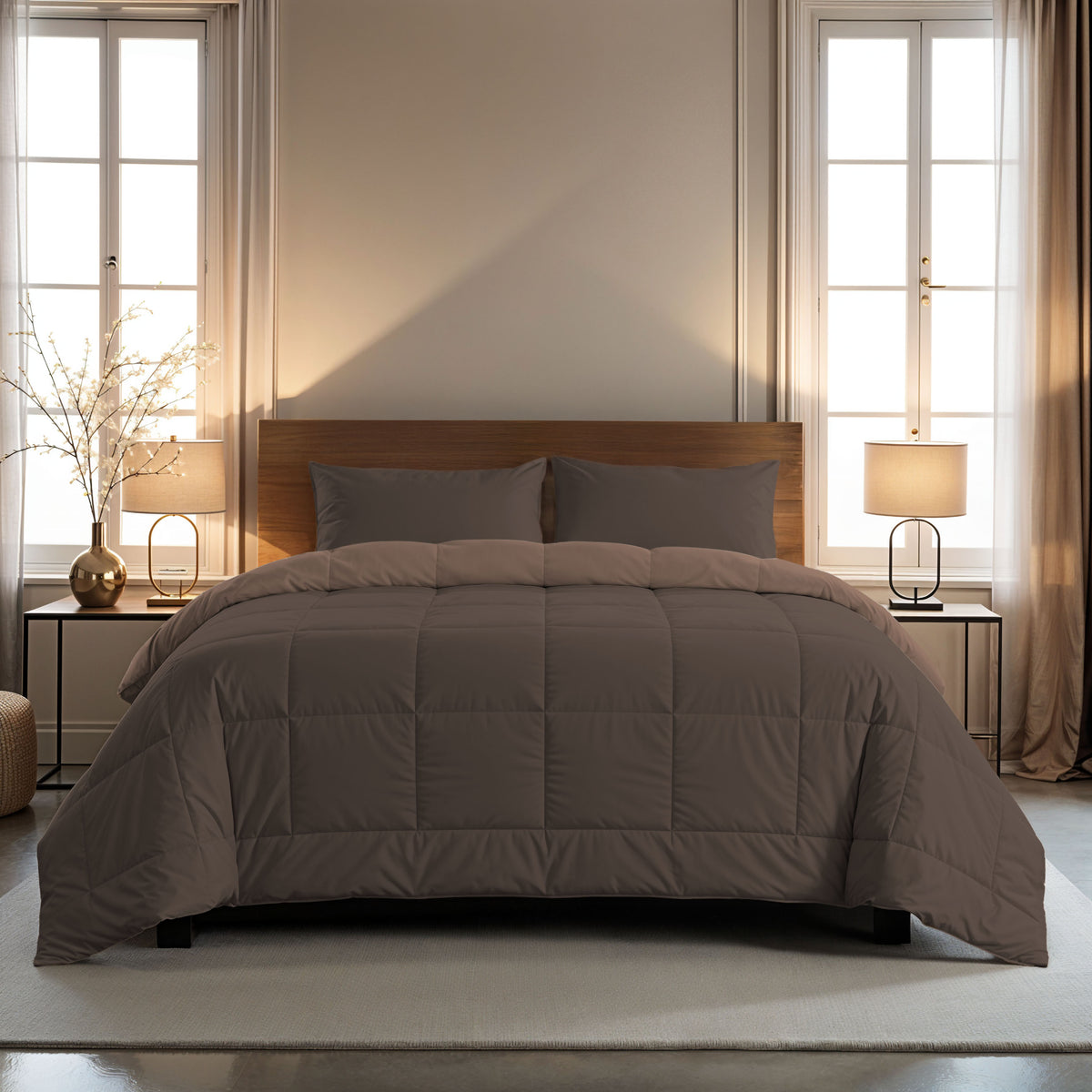 Een keurig opgemaakt bed met het Zelesta Comfort Duo Dekbed in chocolade bruin & mokka en bijpassende kussens staat in het midden tussen twee ramen, geflankeerd door nachtkastjes met lampen in een zacht verlichte slaapkamer.