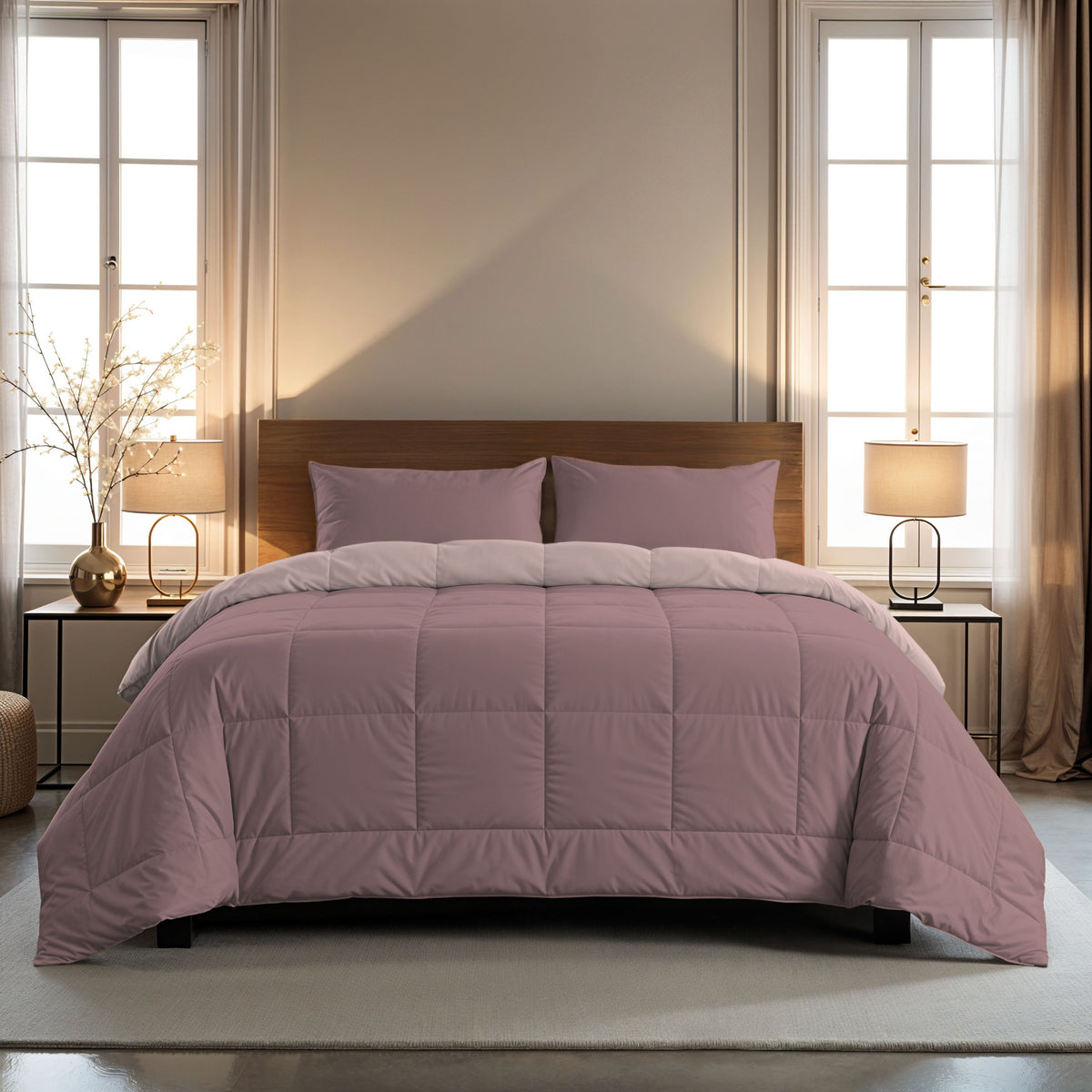 Een netjes opgemaakt bed met het Zelesta Comfort Duo Dekbed in oudroze & lichtroze en bijpassende kussens staat tussen twee ramen, elk met een lamp op een nachtkastje, in een zacht verlichte slaapkamer.
