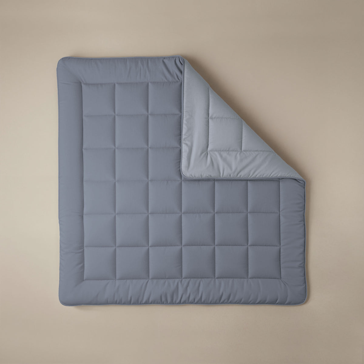 Het Zelesta Comfort Duo Dekbed in ijsblauw en zachtblauw is een netjes opgevouwen, doorgestikte all-season deken met een hoek omgeslagen, uitgestald op een beige ondergrond.