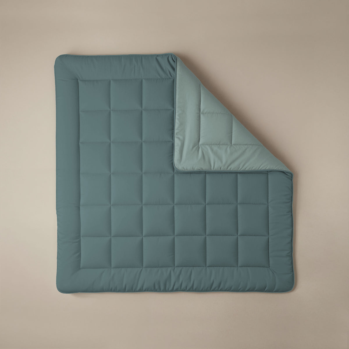 Het Zelesta Comfort Duo Dekbed in mineraal en lichtblauw is plat neergelegd op een beige ondergrond, met een hoek omgeslagen om de lichtblauwe onderkant en de vierkante gewatteerde stiksels te laten zien.