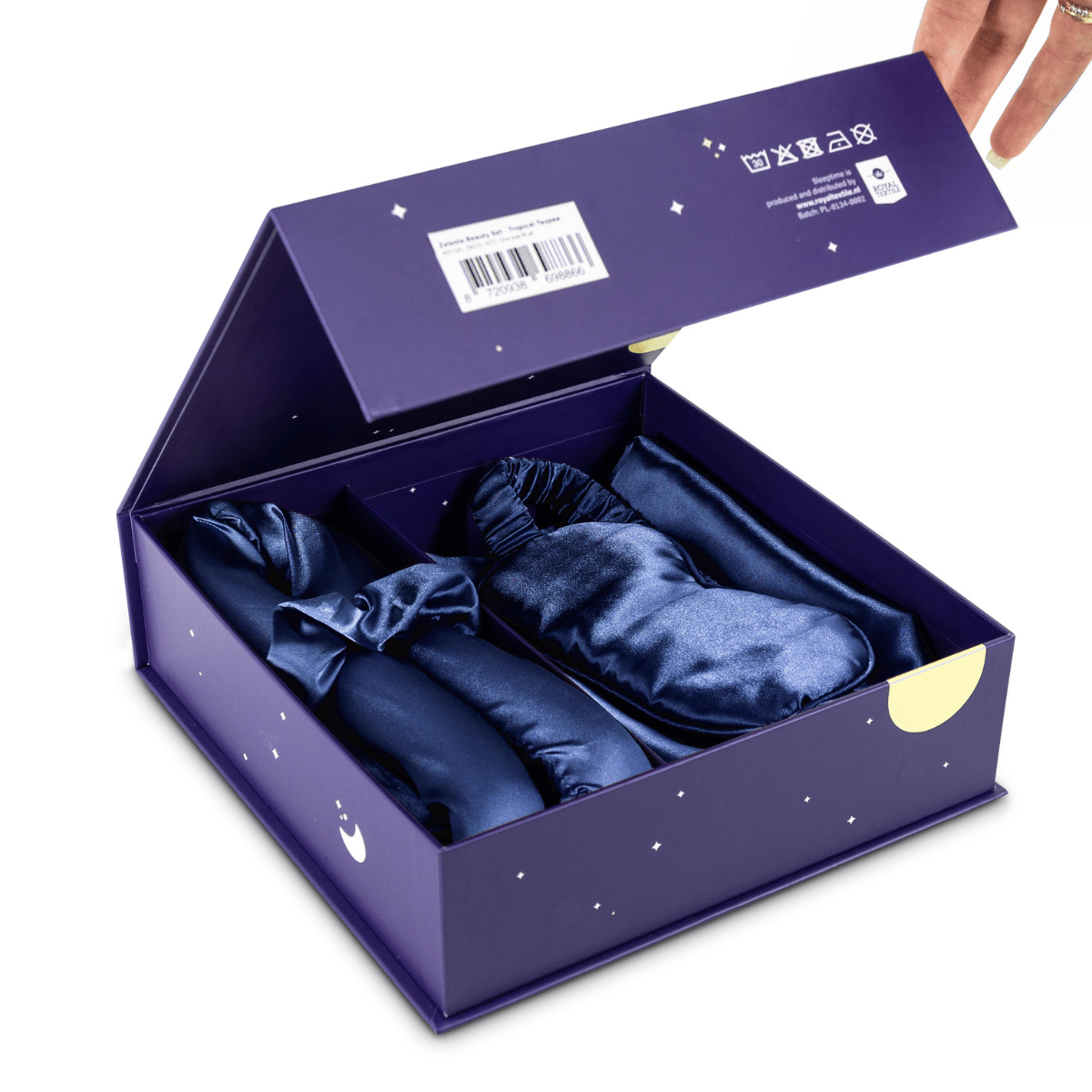 Een hand opent een paarse geschenkdoos om de Zelesta Sleeping Beauty Set - Bliss Blue te onthullen: drie marineblauwe satijnen items, elk in een apart compartiment. Op de doos, die gemaakt is met het oog op anti-veroudering, staan pictogrammen en een streepjescode aan de binnenkant van het deksel.