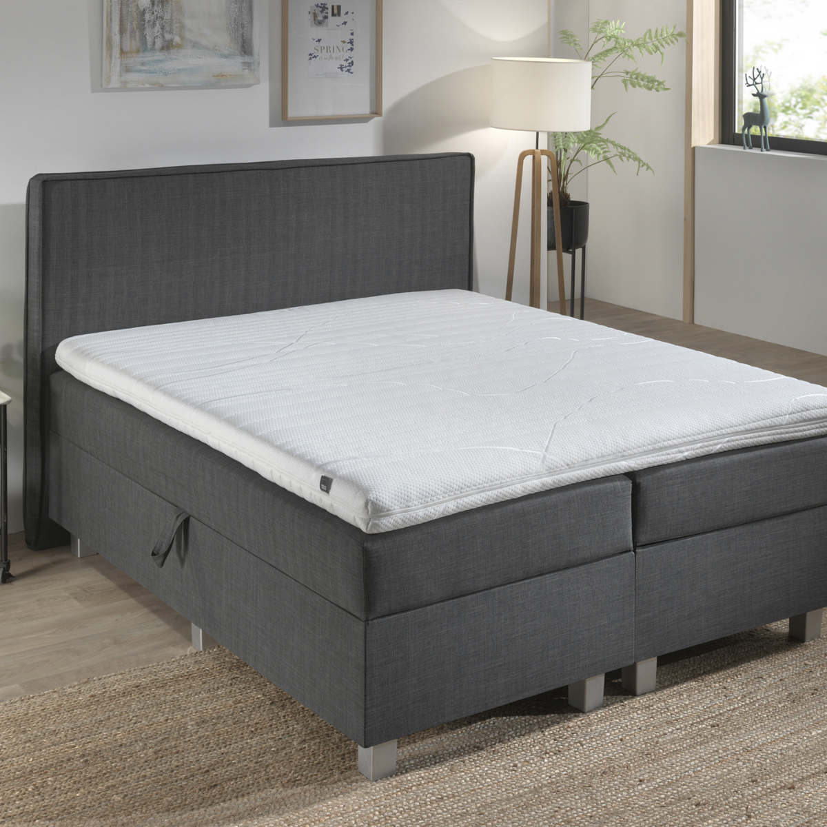 Een moderne slaapkamer met de Silver Line Topper - Hybrid Koudschuim op een grijs gestoffeerd bed, met wit beddengoed, een staande lamp, muurkunst en een geweven vloerkleed van natuurlijke materialen op een houten vloer.