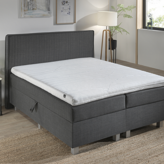 Een moderne slaapkamer met de Silver Line Topper - Hybrid Koudschuim op een grijs gestoffeerd bed, met wit beddengoed, een staande lamp, muurkunst en een geweven vloerkleed van natuurlijke materialen op een houten vloer.