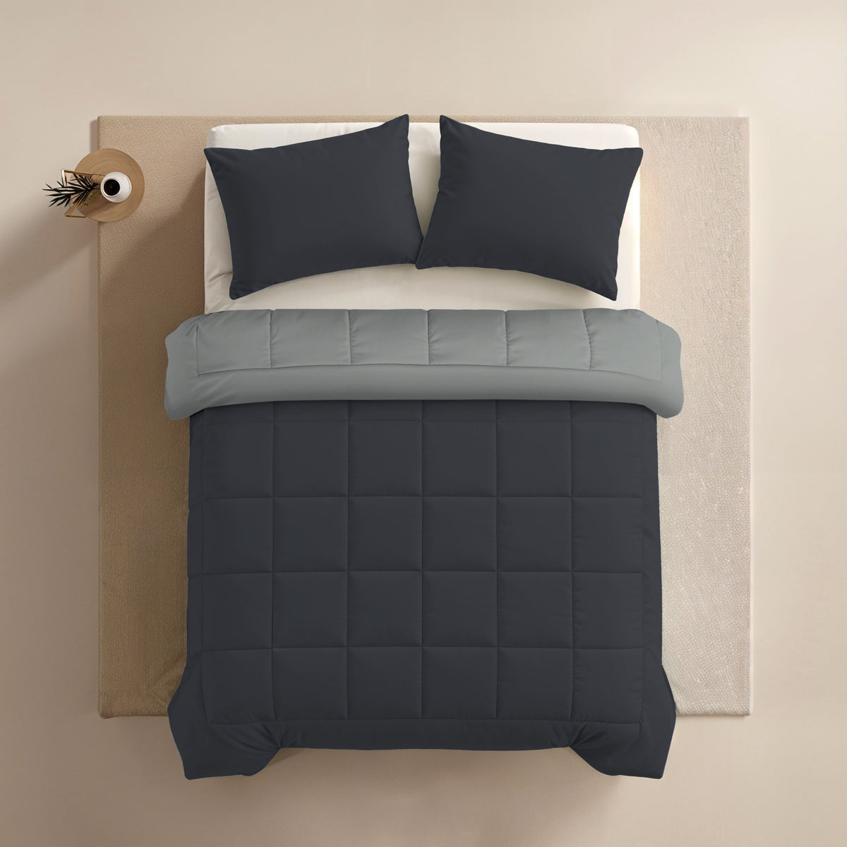 Een keurig opgemaakt bed met luxe Zelesta Comfort Duo Kussenslopen in zacht zwart, een lichtgrijs laken en een donkergrijs gewatteerd dekbed, van bovenaf gezien. Een klein rond nachtkastje met een plant staat links; al het beddengoed is wasmachine geschikt.