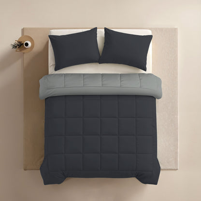Een keurig opgemaakt bed met luxe Zelesta Comfort Duo Kussenslopen in zacht zwart, een lichtgrijs laken en een donkergrijs gewatteerd dekbed, van bovenaf gezien. Een klein rond nachtkastje met een plant staat links; al het beddengoed is wasmachine geschikt.