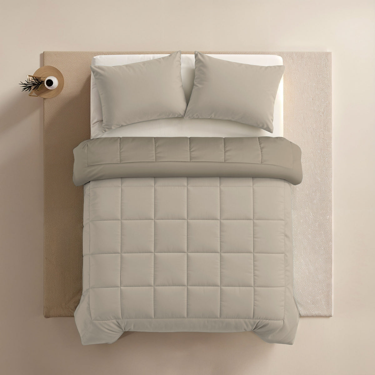 Bovenaanzicht van een netjes opgemaakt bed met het Zelesta Comfort Duo Dekbed in Taupe & Champagne, twee kussens en een klein rond nachtkastje met een enkele tak in een vaas.