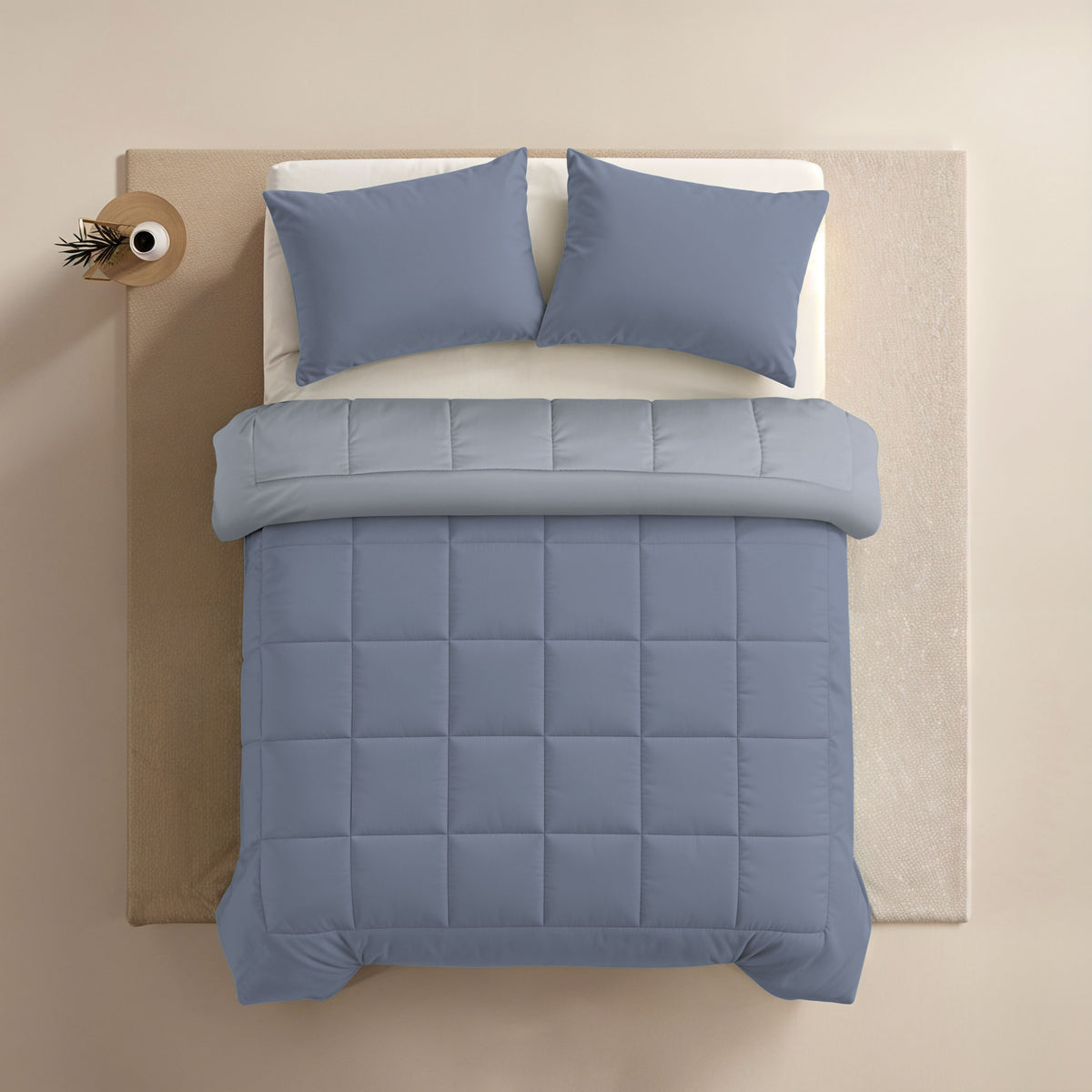Een netjes opgemaakt bed met het Zelesta Comfort Duo Dekbed in ijsblauw & zachtblauw, twee blauwe kussens en een doorgestikt dekbed, van boven gezien op een lichtgekleurd vloerkleed en vloer.