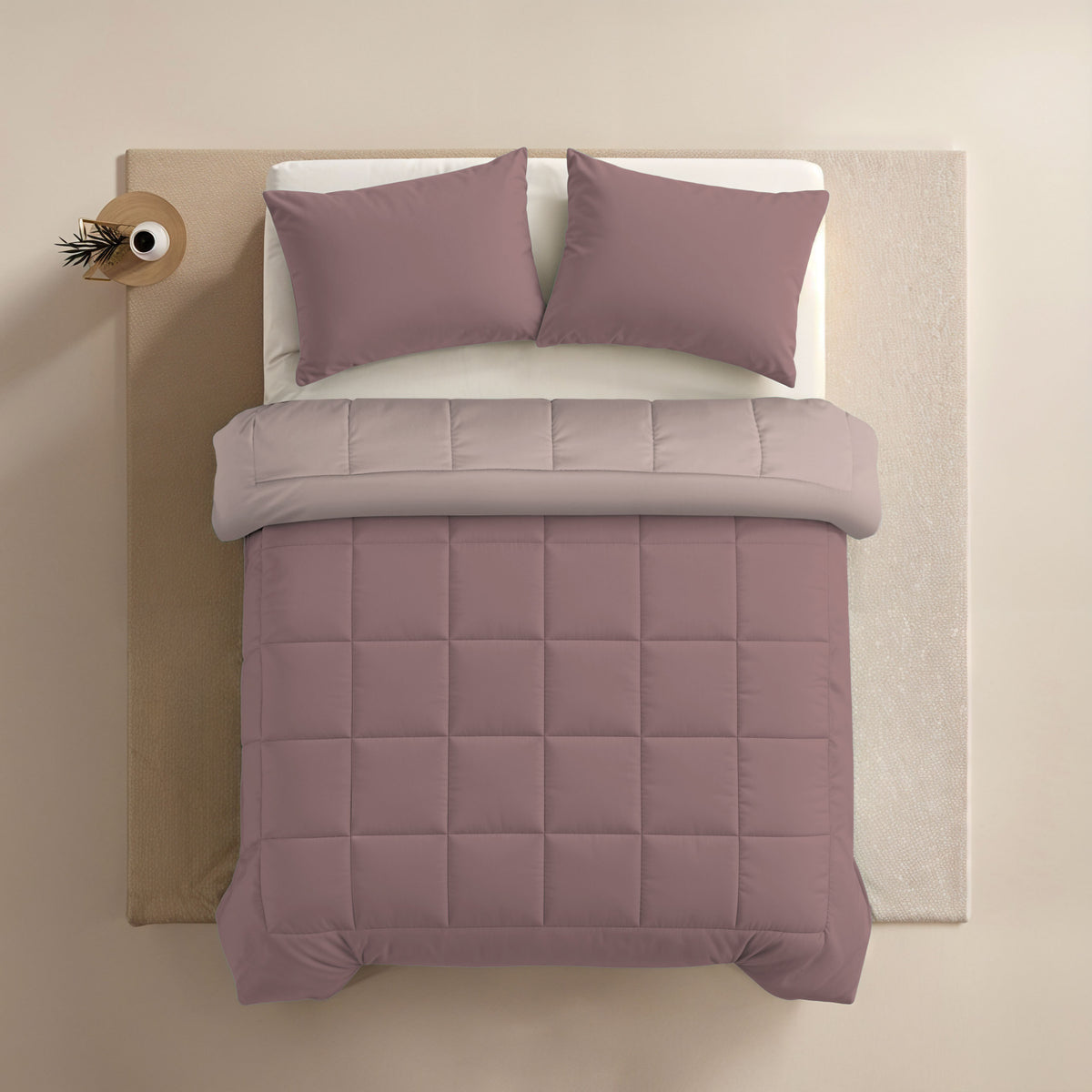 Een netjes opgemaakt bed met het Zelesta Comfort Duo Dekbed in oudroze & lichtroze als een optie voor alle seizoenen, met bijpassende kussens, witte lakens, een beige hoofdeinde en een klein nachtkastje met een vaas met een tak.
