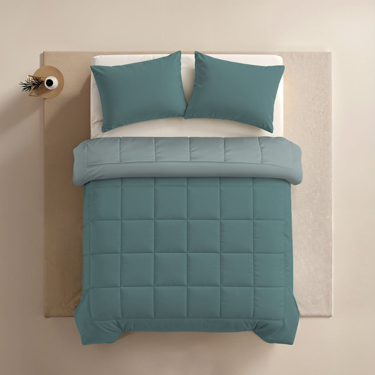Een netjes opgemaakt bed met een groenblauw dekbed, twee Zelesta Comfort Duo Kussenslopen - Mineraal (60x70cm) en een lichtgrijze microvezeldeken op beige, aangevuld met een klein rond bijzettafeltje met een plant.