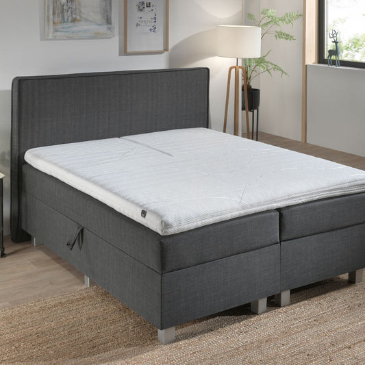 Een moderne slaapkamer heeft een grijs gestoffeerd bed met stevige ondersteuning en de Silver Line Split Topper - Hybrid Koudschuim, houten vloer van natuurlijke materialen, geweven vloerkleed, staande lamp en ingelijste muurkunst voor een gezonde slaapomgeving.