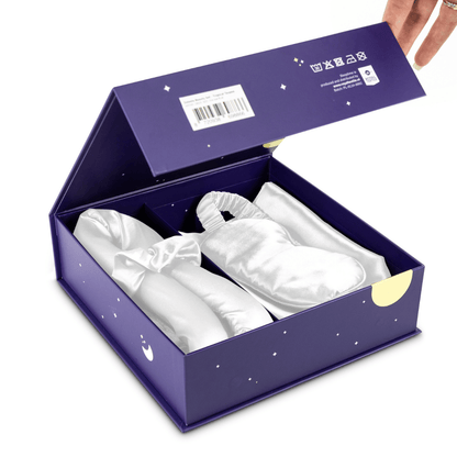 Een hand opent de Zelesta Sleeping Beauty Set - Wonderful White, en onthult elegante witte satijnen pantoffels en een bijpassend Mulberry zijden slaapmasker in een paarse doos met sterrendessins voor een anti-aging vleugje luxe.