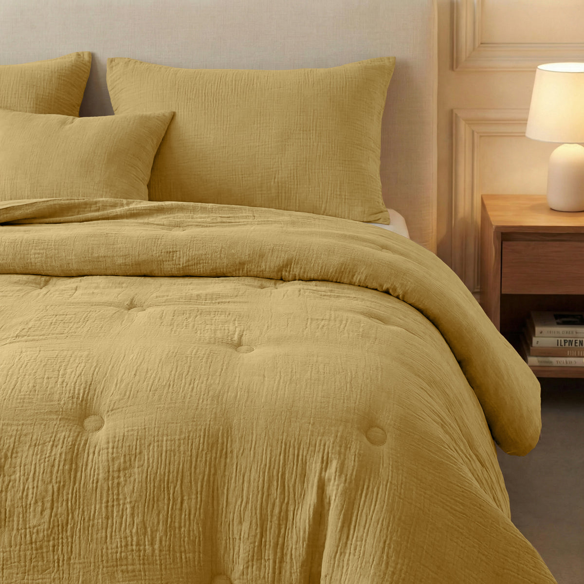 Een keurig opgemaakt bed met de Zelesta Japandibed Katoen Mousseline in oker, gestyled met kussens bij een houten nachtkastje met een lamp en gestapelde boeken, tegen een beige muur.