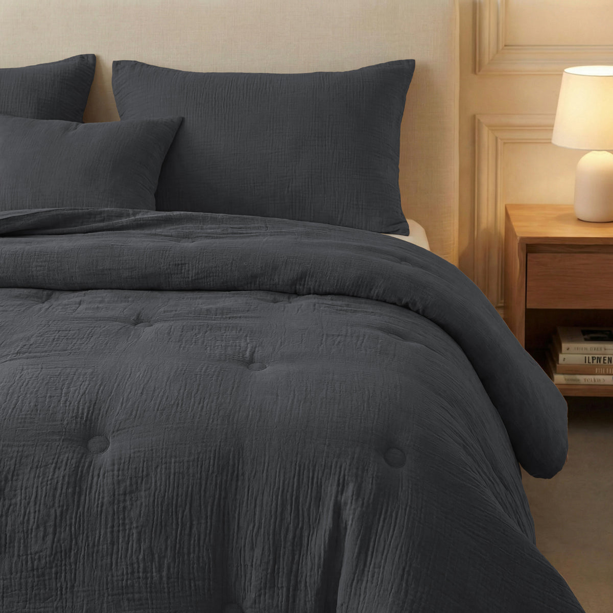 Een keurig opgemaakt bed met de Zelesta Japandibed Katoen Mousseline in antraciet, bijpassende kussens, een beige hoofdeinde en een houten nachtkastje met daarop een lamp en gestapelde boeken.