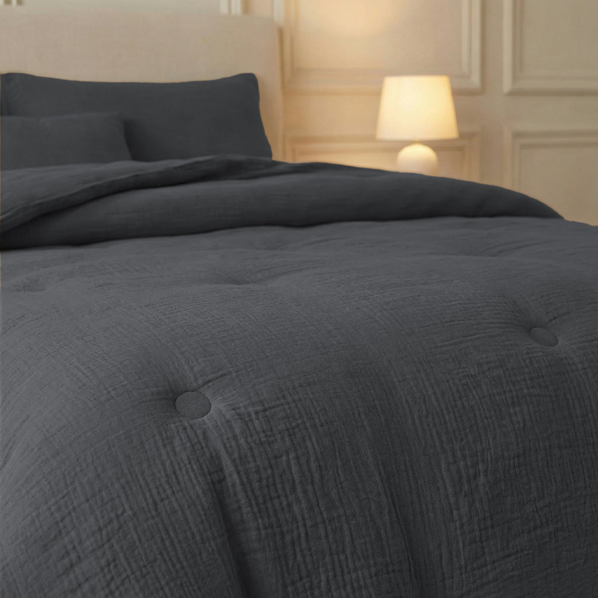 Een netjes opgemaakt bed met de Zelesta Japandibed Katoen Mousseline in antraciet en bijpassende kussens rust tegen een beige hoofdeinde. Een brandende tafellamp staat op het nachtkastje in een zacht verlichte, crèmekleurige kamer.