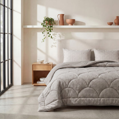 Een netjes opgemaakt bed met het Zelesta Flanel Luxe - Taupe & Grijs temperatuurregulerend dekbed staat naast een houten nachtkastje en een plank met keramiek decor en een potplant in een lichte, minimalistische slaapkamer.