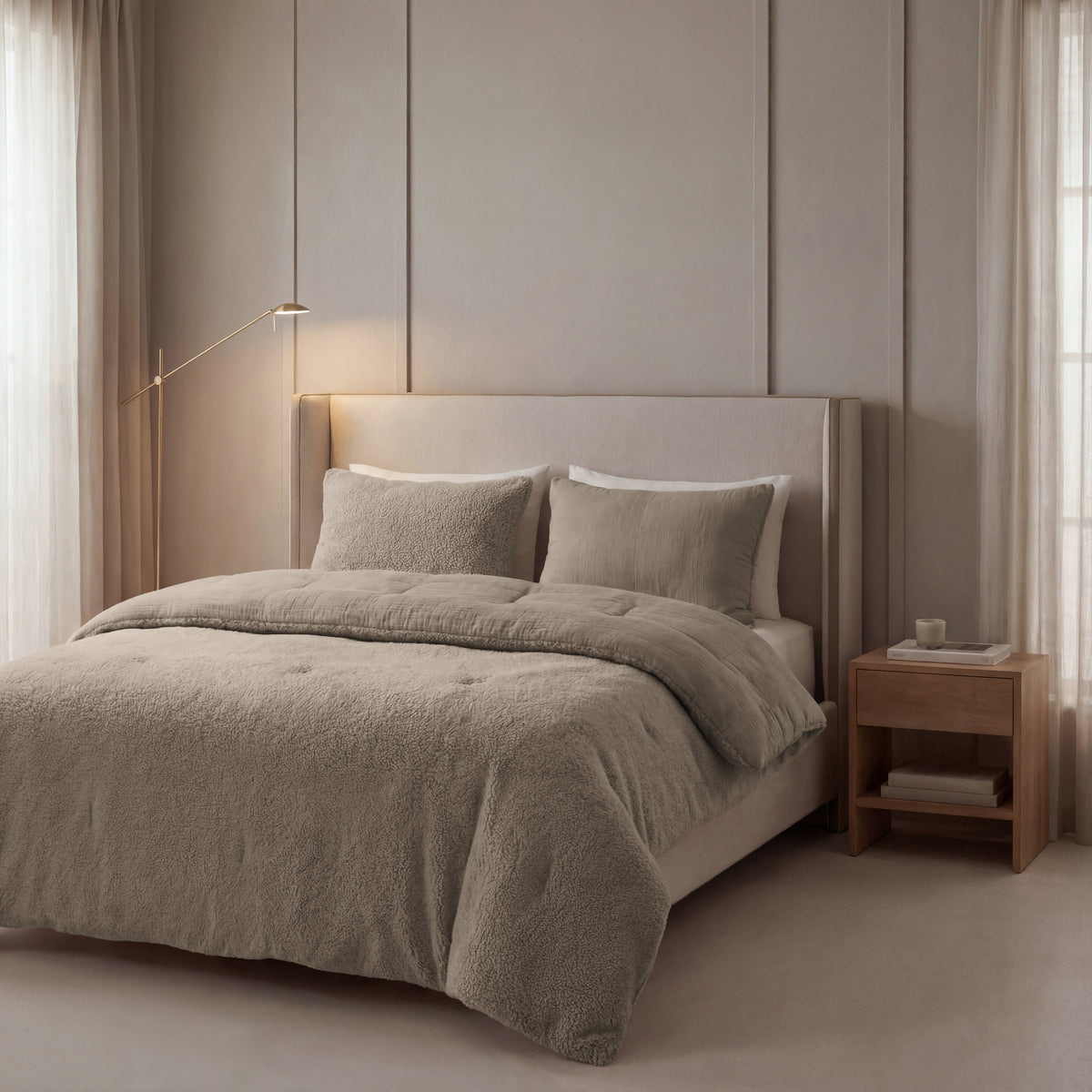 Een moderne, neutraal getinte slaapkamer met een groot gestoffeerd bed met beddengoed van Zelesta Teddy Katoen Mousseline - Taupe, een houten nachtkastje, minimalistische inrichting, natuurlijk licht en een zacht gloeiende vloerlamp.