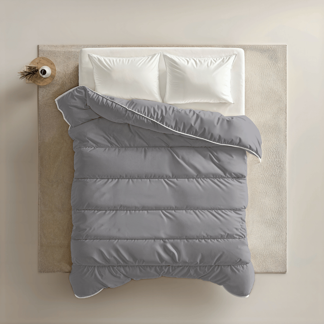 Een netjes opgemaakt bed met twee witte kussens en een grijs Zelesta Royalbed - Antraciet, van bovenaf gezien, geplaatst op een lichtbeige vloerkleed met links een klein rond nachtkastje.