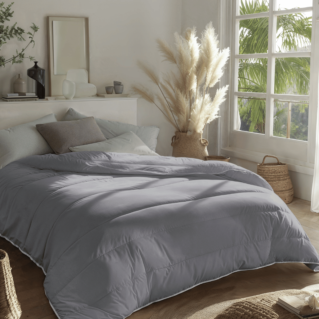 Een netjes opgemaakt bed met een donkergrijs Zelesta Royalbed - Antraciet dekbed in een lichte slaapkamer, ingericht met neutrale kussens, rieten manden en pampagras bij een groot raam.