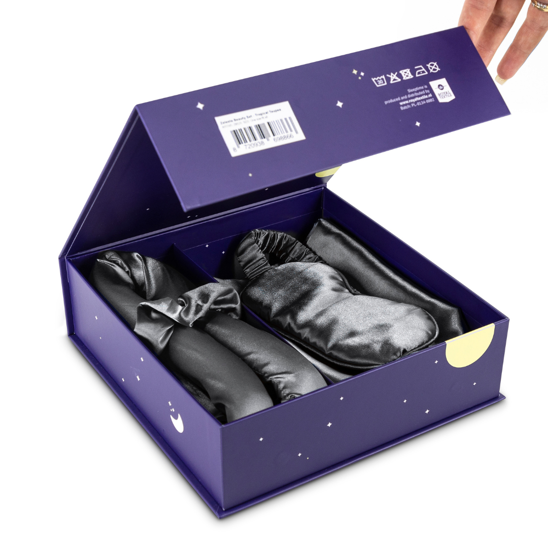 Een hand opent de Zelesta Sleeping Beauty Set - Amazing Anthracite en onthult drie zwart satijnen slaapaccessoires, waaronder een anti-aging slaapmasker en een luxe Mulberry zijden kussensloop.