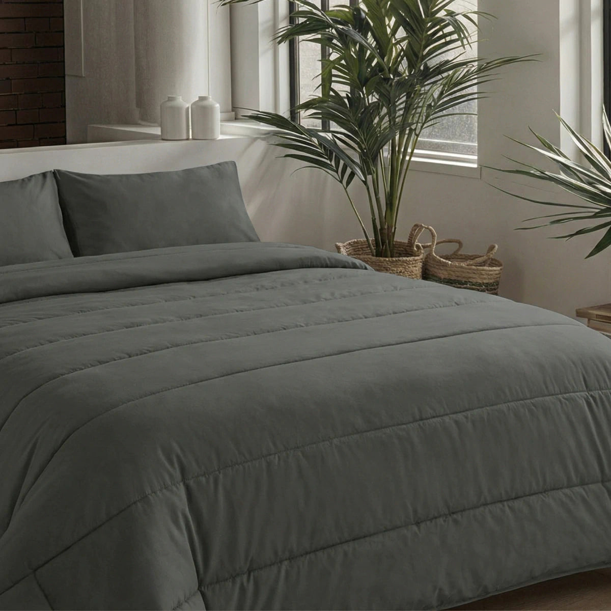 Een moderne slaapkamer heeft een netjes opgemaakt bed met het Zelesta Comfort Uni Dekbed in antraciet en twee kussens, bij grote ramen omringd door potplanten en decoratieve manden.