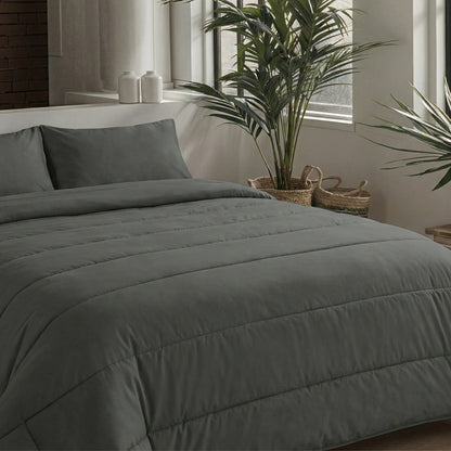 Een moderne slaapkamer heeft een netjes opgemaakt bed met het Zelesta Comfort Uni Dekbed in antraciet en twee kussens, bij grote ramen omringd door potplanten en decoratieve manden.