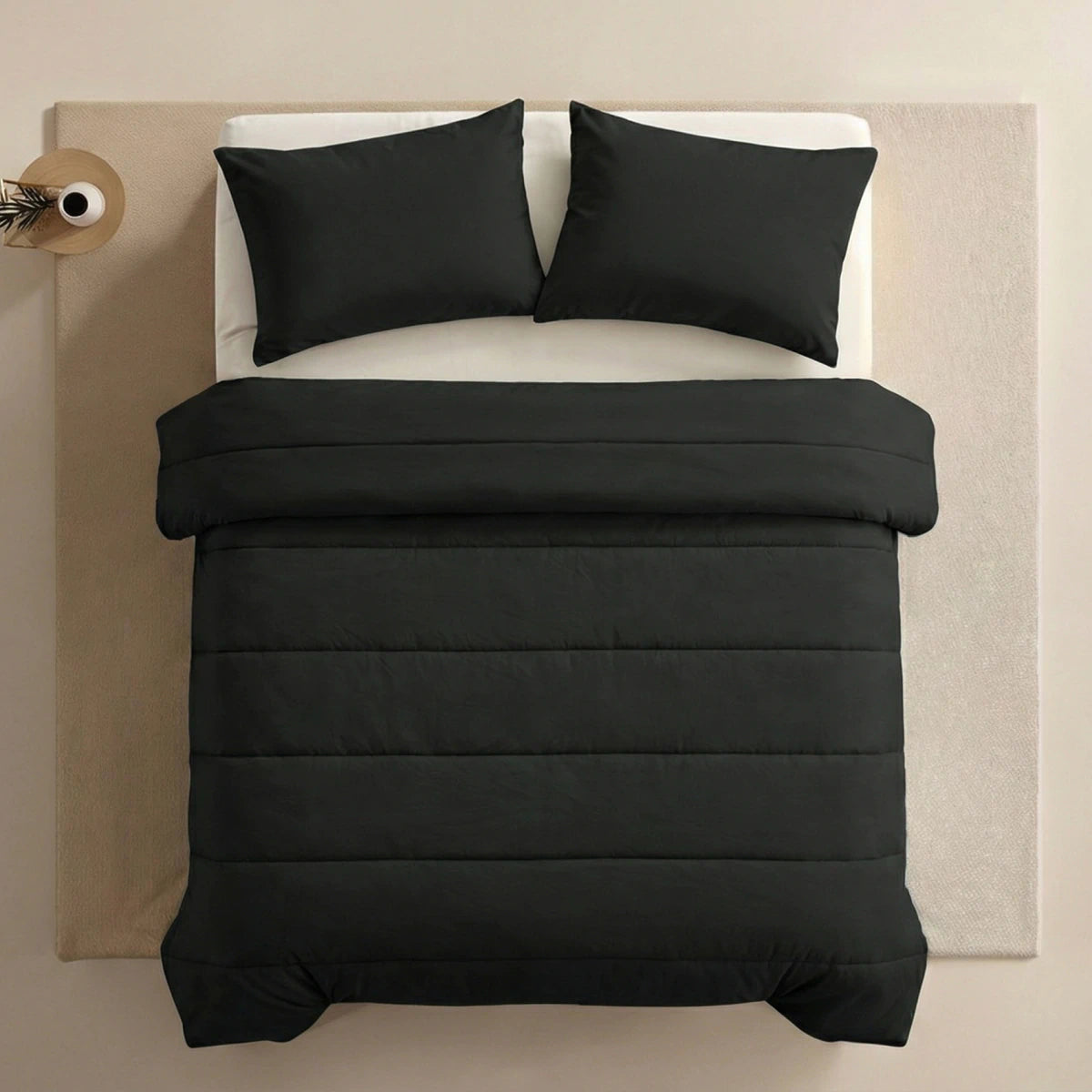 Een netjes opgemaakt bed met het Zelesta Comfort Uni Dekbed - Zwart en bijpassende zwarte kussens, op een beige vloerkleed naast een ronde bijzettafel met een vaas.