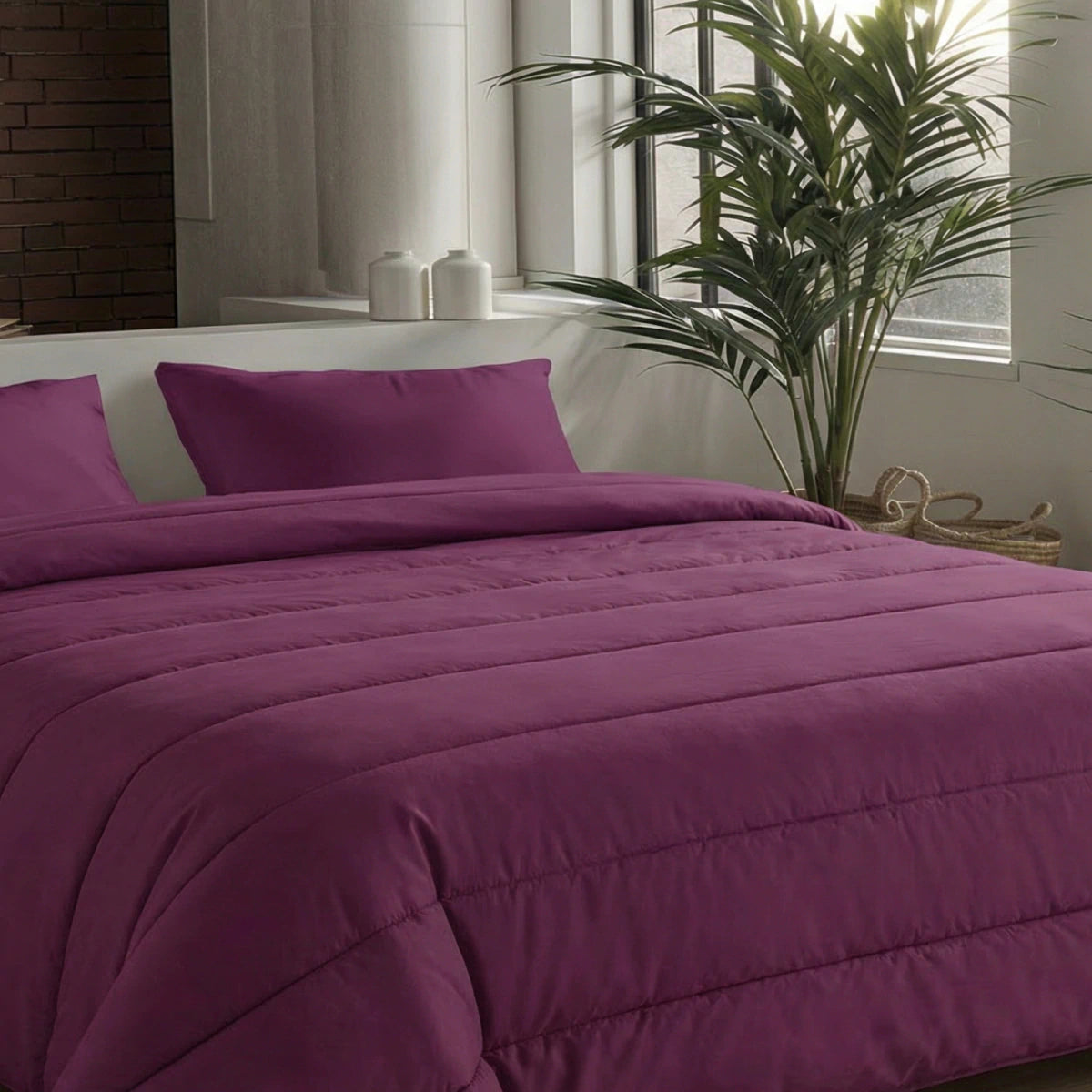 Een keurig opgemaakt bed met het Zelesta Comfort Uni Dekbed in Bordeaux en bijpassende kussens staat naast een potplant en een raam gedrapeerd met witte gordijnen.