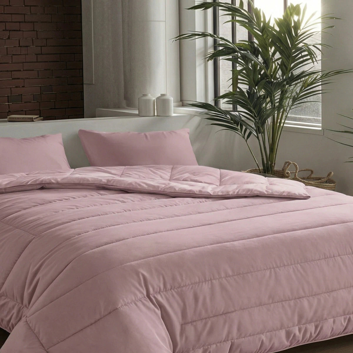 Een netjes opgemaakt bed met het Zelesta Comfort Uni Dekbed - Oud Roze en bijpassende kussens in een moderne slaapkamer met grote ramen, een potplant en een bakstenen accentmuur - ideaal voor klassieke dekbedden of als dekbed voor het hele seizoen.