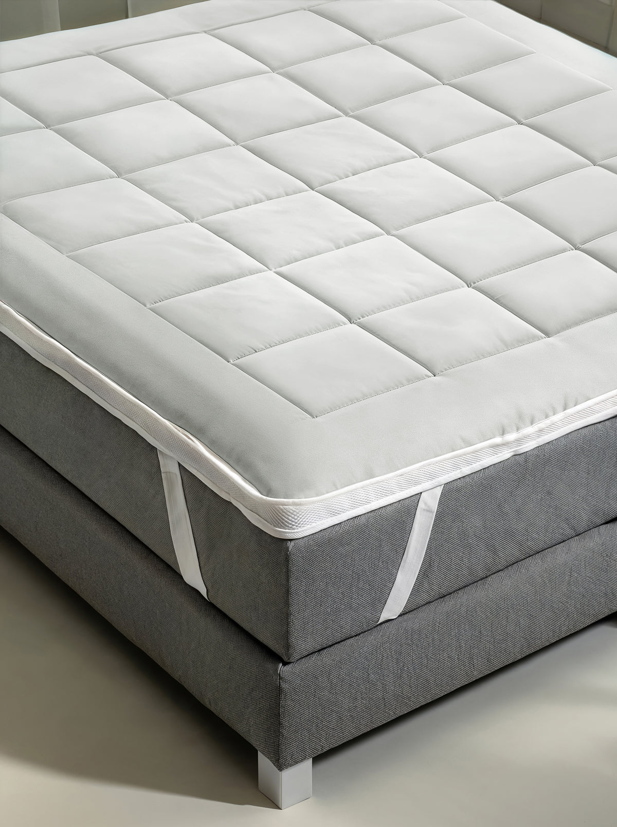 De Zelesta Topper, een witte gewatteerde matras topper met elastische hoekbanden, wordt geplaatst op een grijze bedbodem met metalen poten, wat zorgt voor meer slaapcomfort en ondersteuning.