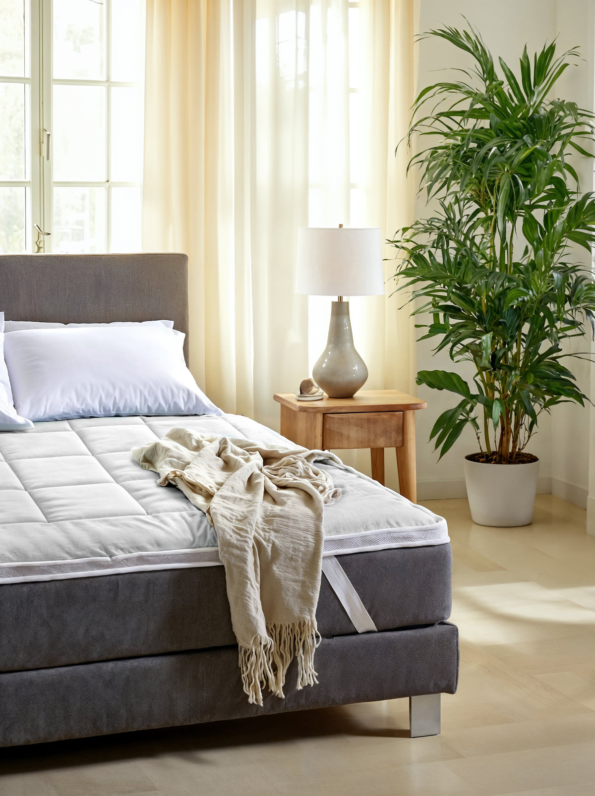 Een netjes opgemaakt bed met een wit kussen en een beige deken naast een houten nachtkastje, een lamp en een grote potplant tonen slaapcomfort versterkt door de Zelesta Topper in een lichte, zonovergoten slaapkamer.