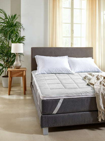 Een keurig opgemaakt bed met witte kussens en een Zelesta Topper met Blue Cell Technologie staat naast een houten nachtkastje met een lamp en plant, in een lichte kamer met lichte gordijnen voor optimaal slaapcomfort.