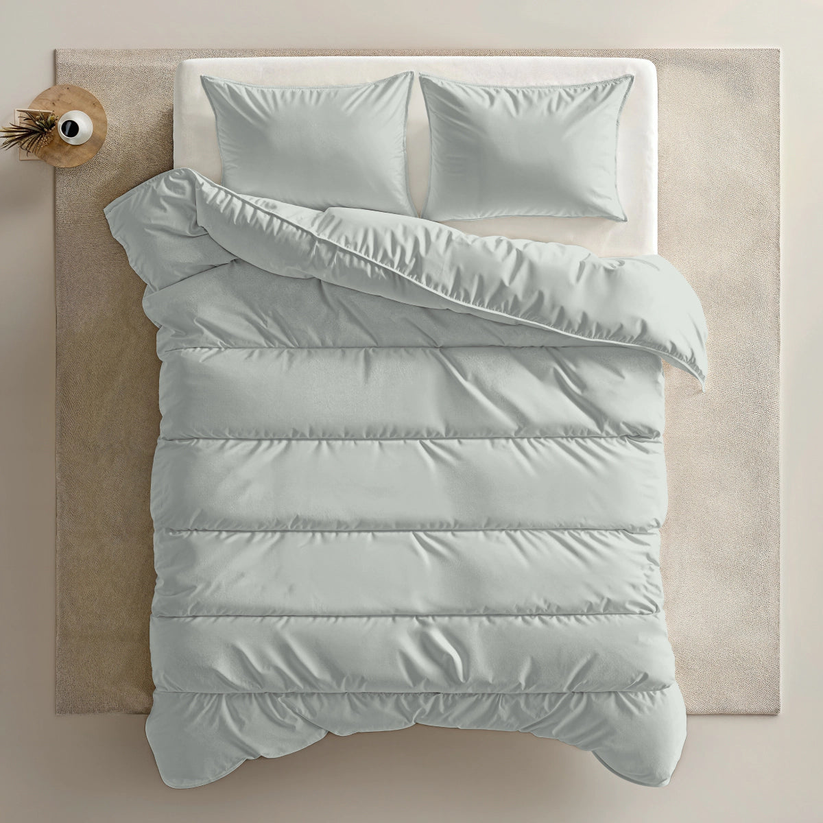 Bovenaanzicht van een netjes opgemaakt Zelesta Everyday Uni Dekbed - Mistgroen met lichtgrijs beddengoed en twee kussens, op een beige vloerkleed naast een ronde houten tafel met een vaas-dekbed en overtrek in één voor comfort het hele jaar door.