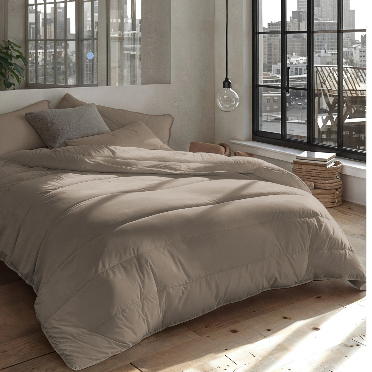 Een netjes opgemaakt bed met een Zelesta Everyday Uni Dekbed - Zachtbruin staat bij grote ramen met uitzicht op een stadslandschap, in een kamer met houten vloeren, neutrale muren en een hangende lamp.