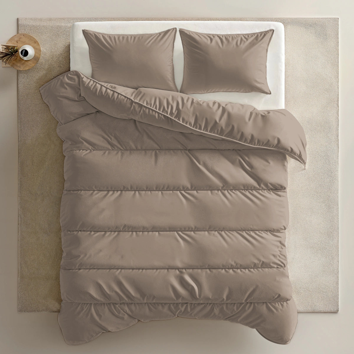 Een keurig opgemaakt bed met een Zelesta Everyday Uni Dekbed - Zachtbruin en twee kussens, op een lichtgekleurd vloerkleed naast een klein rond bijzettafeltje met een plant.
