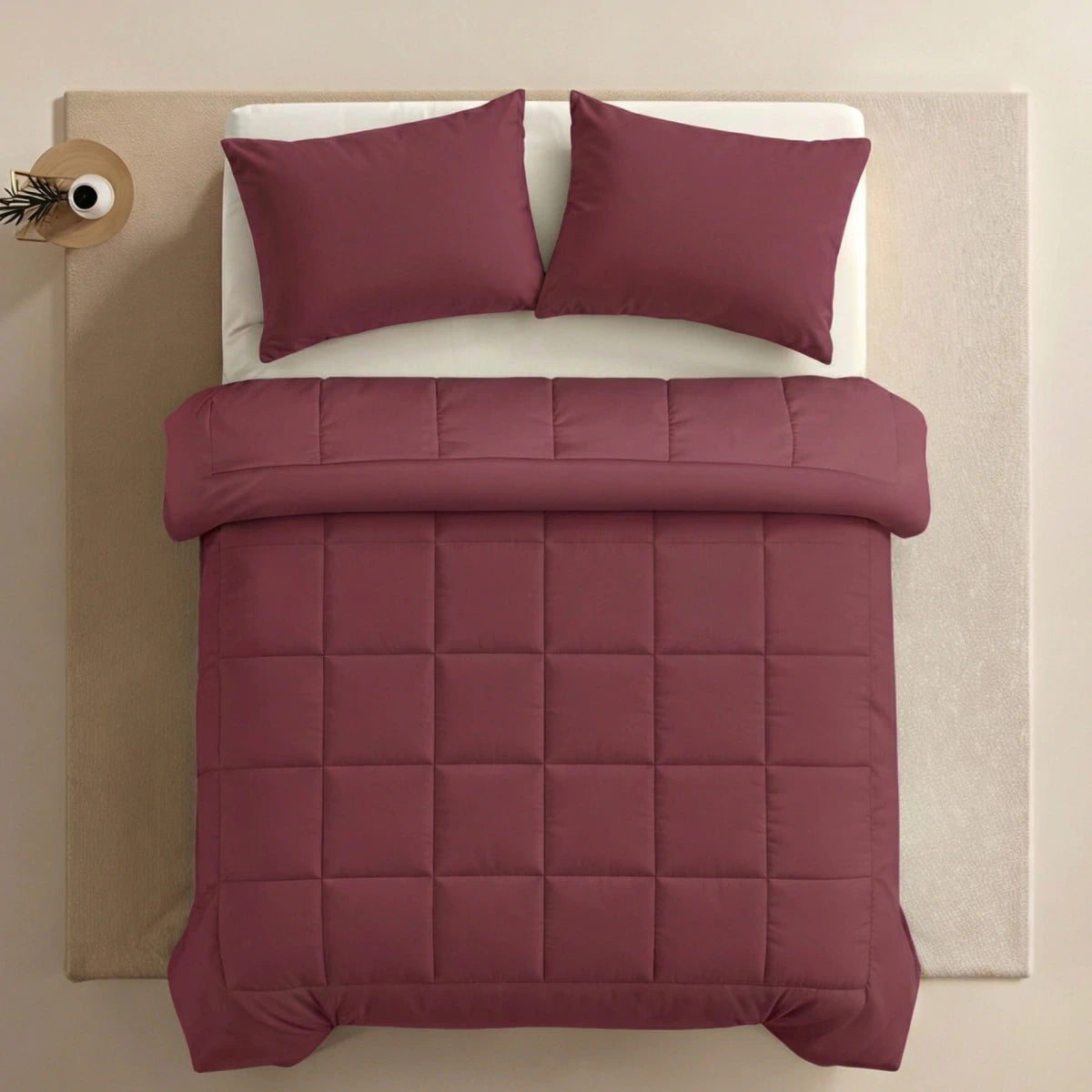 Een keurig opgemaakt bed met een Zelesta Pure Uni Dekbed Katoen in bordeaux rood en twee bijpassende kussens, op een beige vloerkleed naast een klein rond bijzettafeltje met een enkele vaas.