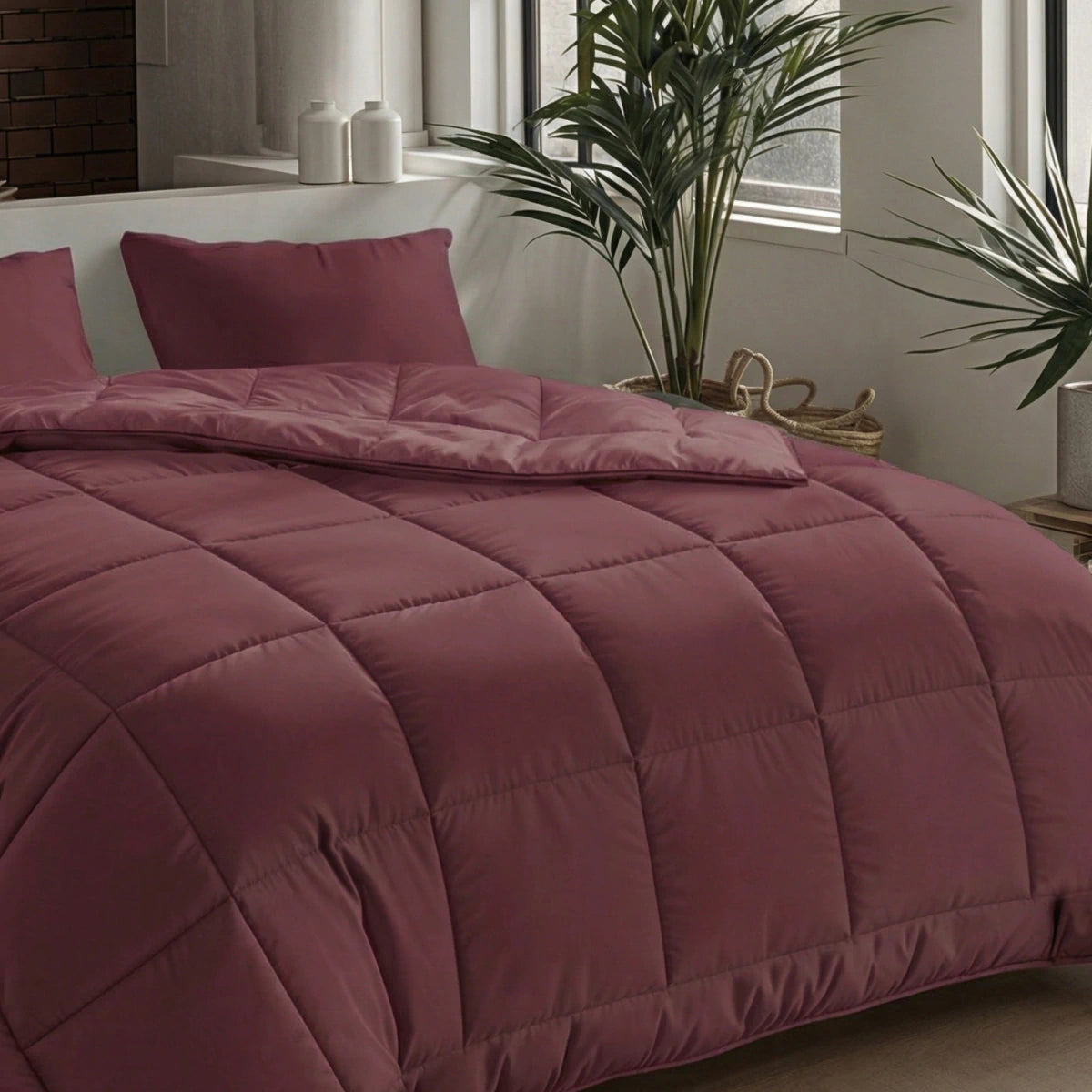Een netjes opgemaakt bed met het Zelesta Pure Uni Dekbed Katoen in bordeaux rood en kussens, bij een raam met potplanten en neutrale inrichting, brengt een ademend slaapcomfort in deze moderne slaapkamer.