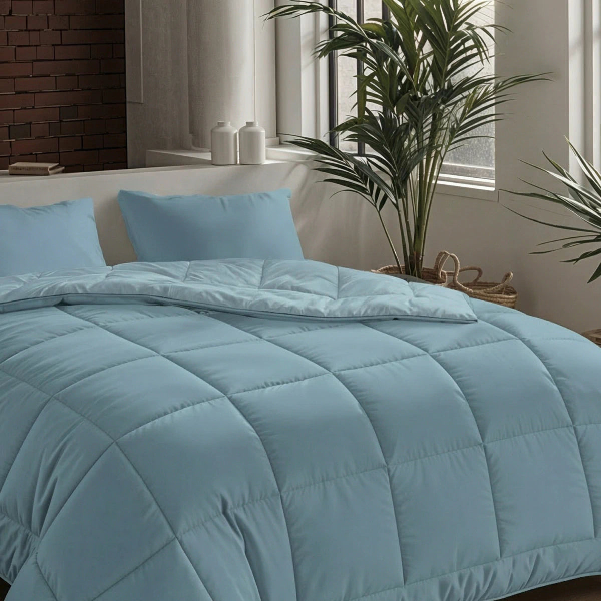 Een netjes opgemaakt bed met de Zelesta Pure Uni Dekbed Katoen in sereen blauw en twee bijpassende kussens, geplaatst bij grote ramen en kamerplanten voor een ademend slaapcomfort in een moderne slaapkamer.