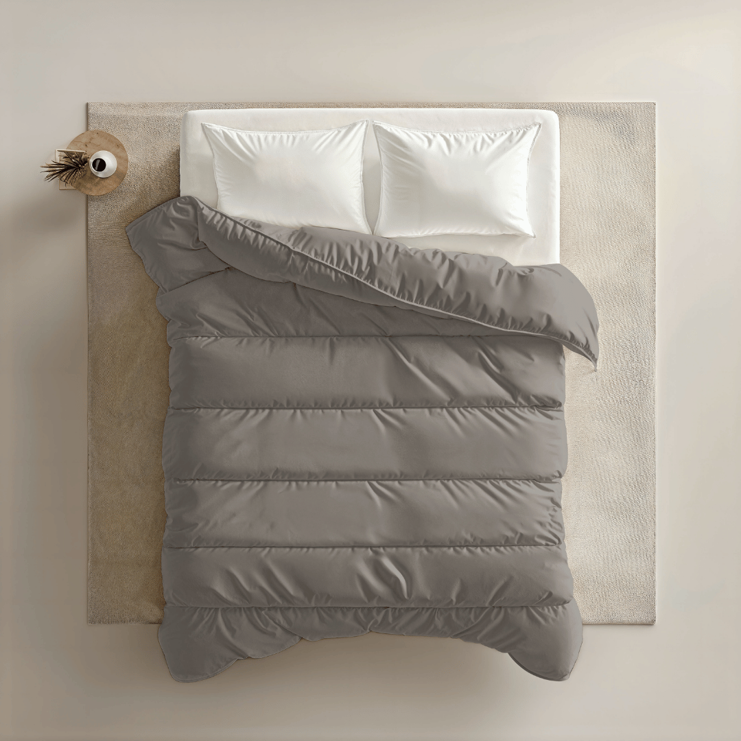 Bovenaanzicht van een netjes opgemaakt bed met het Zelesta Royalbed - Tender Grey met twee witte kussens, een grijs dekbed en een beige vloerkleed, naast een rond houten nachtkastje met daarop een decoratief item.