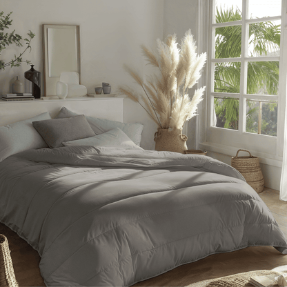 Een netjes opgemaakt bed met Zelesta Royalbed - Tender Grey beddengoed biedt het hele seizoen comfort in een zonovergoten kamer met grote ramen, pampagras in een vaas en een rustgevende neutrale inrichting.