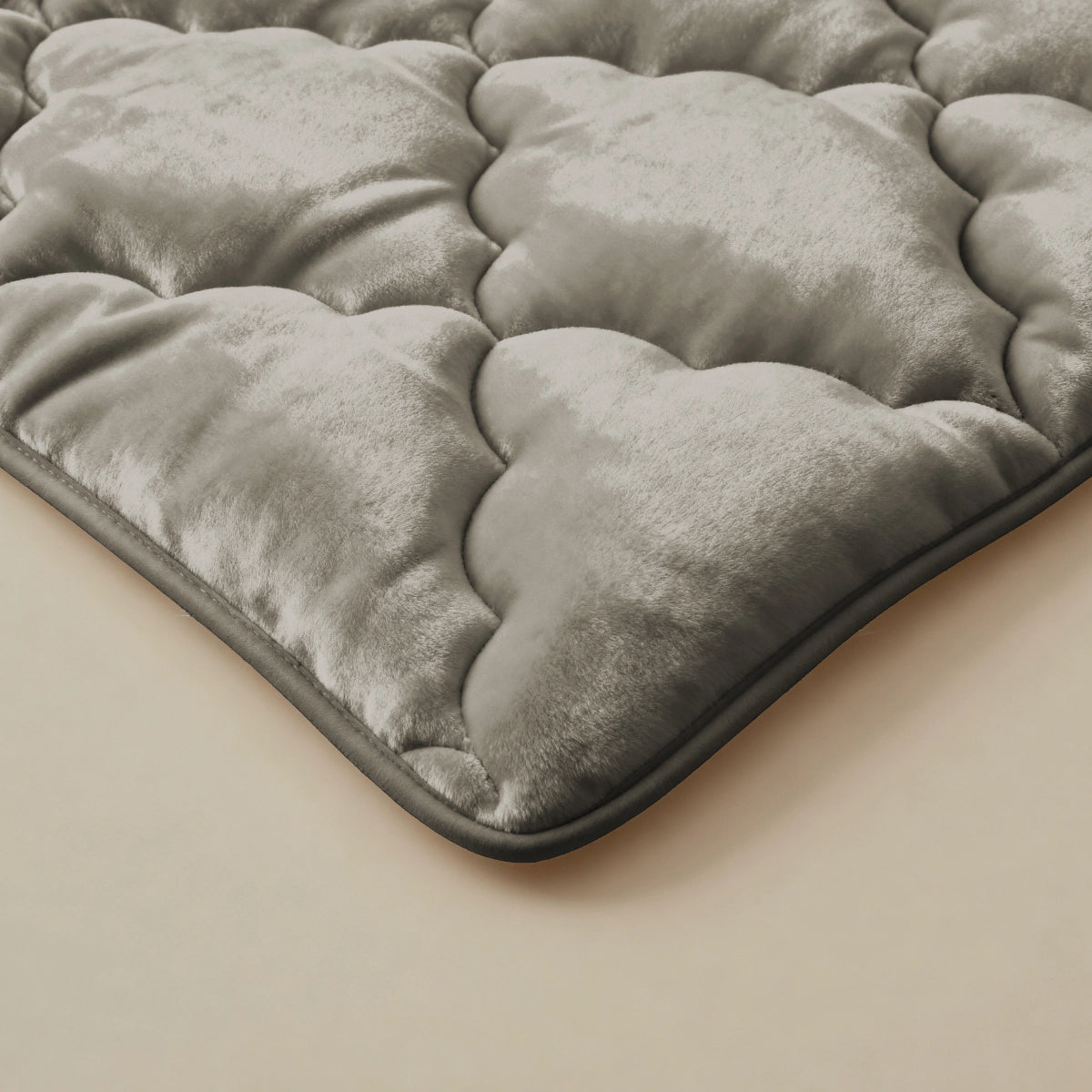 Een close-up van de Zelesta Flanel Uni - Taupe toont de zachte, taupe, gewatteerde textuur en het pluche materiaal - vergelijkbaar met een temperatuurregulerend 2-in-1 dekbed - rustend op een beige oppervlak met een lichte glans en zacht gewatteerde delen.