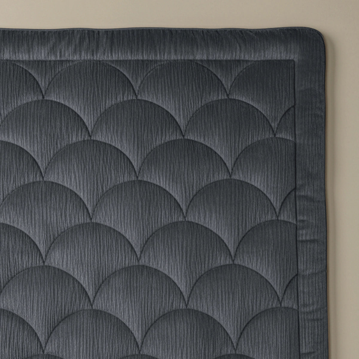 Het antracietkleurige Zelesta Flanel Velvetbed Chique met schulpmotief en brede rand wordt gedeeltelijk getoond op een beige oppervlak, waarbij het gestructureerde, gecapitonneerde ontwerp zichtbaar is.