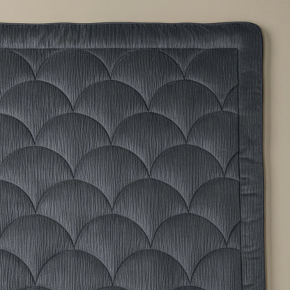 Het antracietkleurige Zelesta Flanel Velvetbed Chique met schulpmotief en brede rand wordt gedeeltelijk getoond op een beige oppervlak, waarbij het gestructureerde, gecapitonneerde ontwerp zichtbaar is.