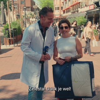 Rob Geus in witte doktersjas interviewt tevreden klant met Zelesta dekbed op bruisend plein