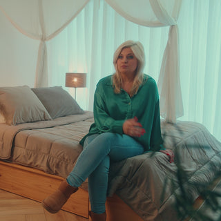 Bobby Eden zit op bed met Zelesta dekbed in beige – draagt groene blouse en praat in sfeervolle slaapkamer met witte canopy-gordijnen